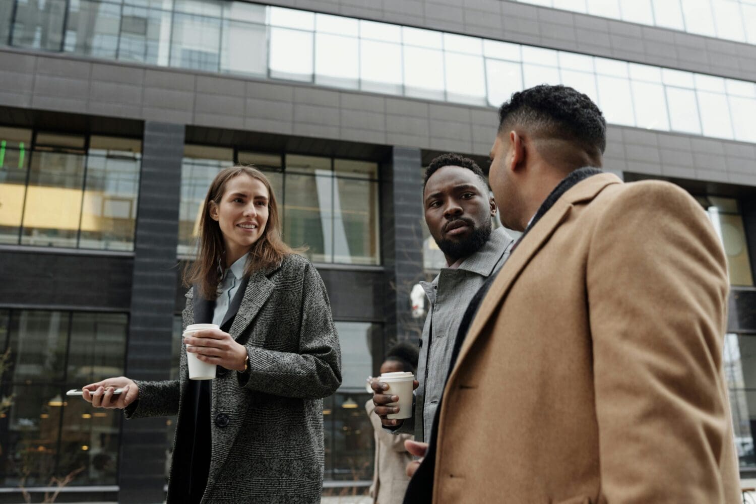 Three people in professional attire talk over coffee outside an office building, illustrating how to ask MBA alumni about their career growth and challenges.