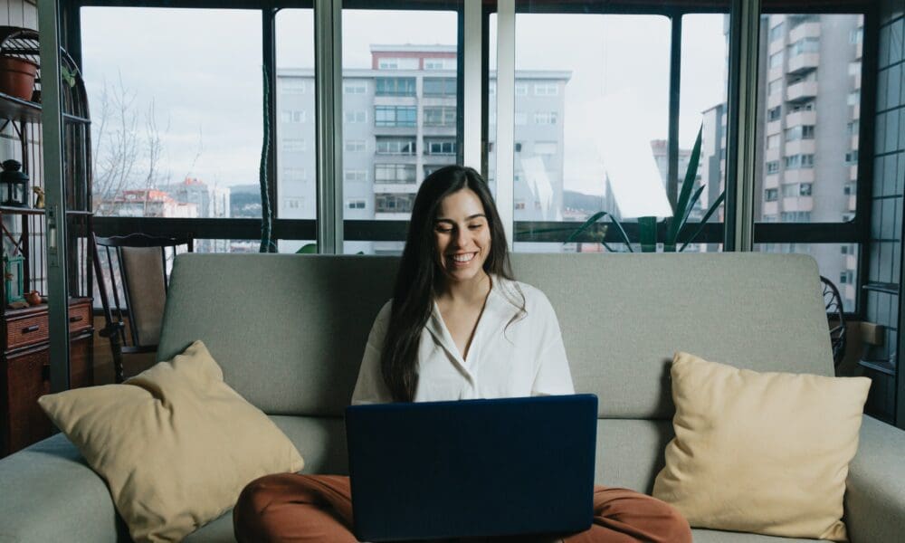Woman researching MBA school selection options on laptop while sitting on couch in bright urban apartment.